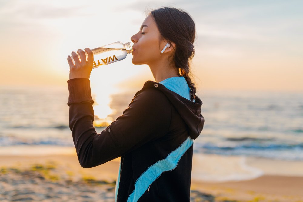 attractive slim woman doing sport exercises morning sunrise beach sports wear thirsty drinking water bottle healthy lifestyle listening music wireless earphones smiling happy